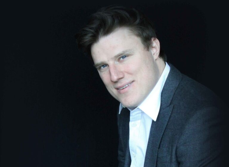 Young man with light brown hair and blue eyes, wearing a grey blazer and white shirt, smiling slightly against a dark background.